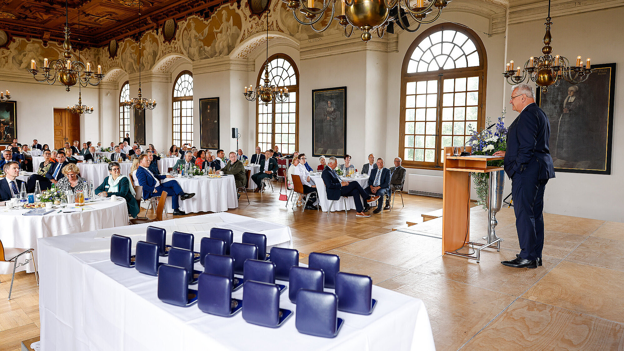 Blick in den Festsaal, rechts Innenminister Herrmann am Rednerpult, links Publikum in Stuhlreihen, im Vordergrund kommunale Verdienstmedaillen auf Tisch in Schachteln