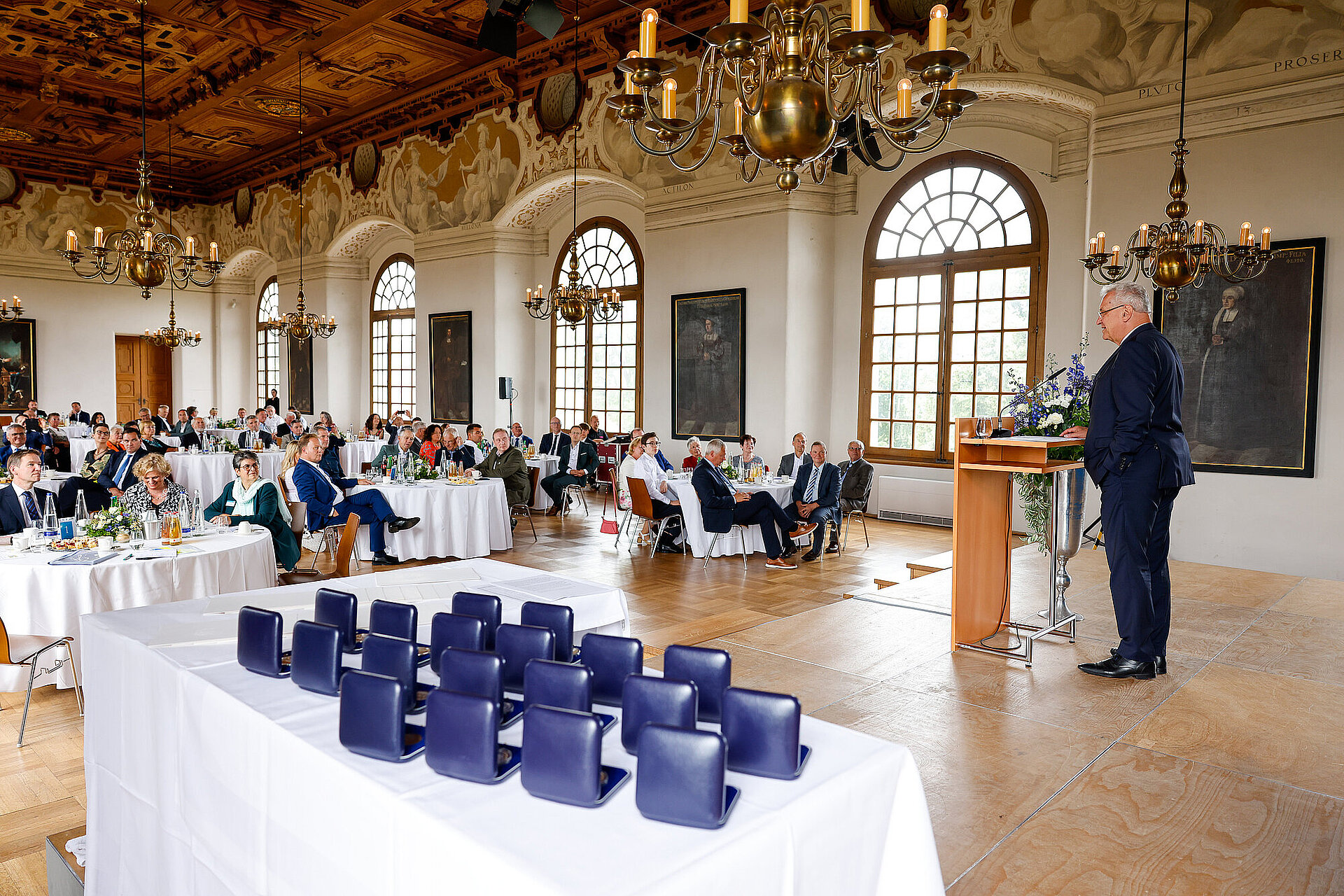 Blick in den Festsaal, rechts Innenminister Herrmann am Rednerpult, links Publikum in Stuhlreihen, im Vordergrund kommunale Verdienstmedaillen auf Tisch in Schachteln