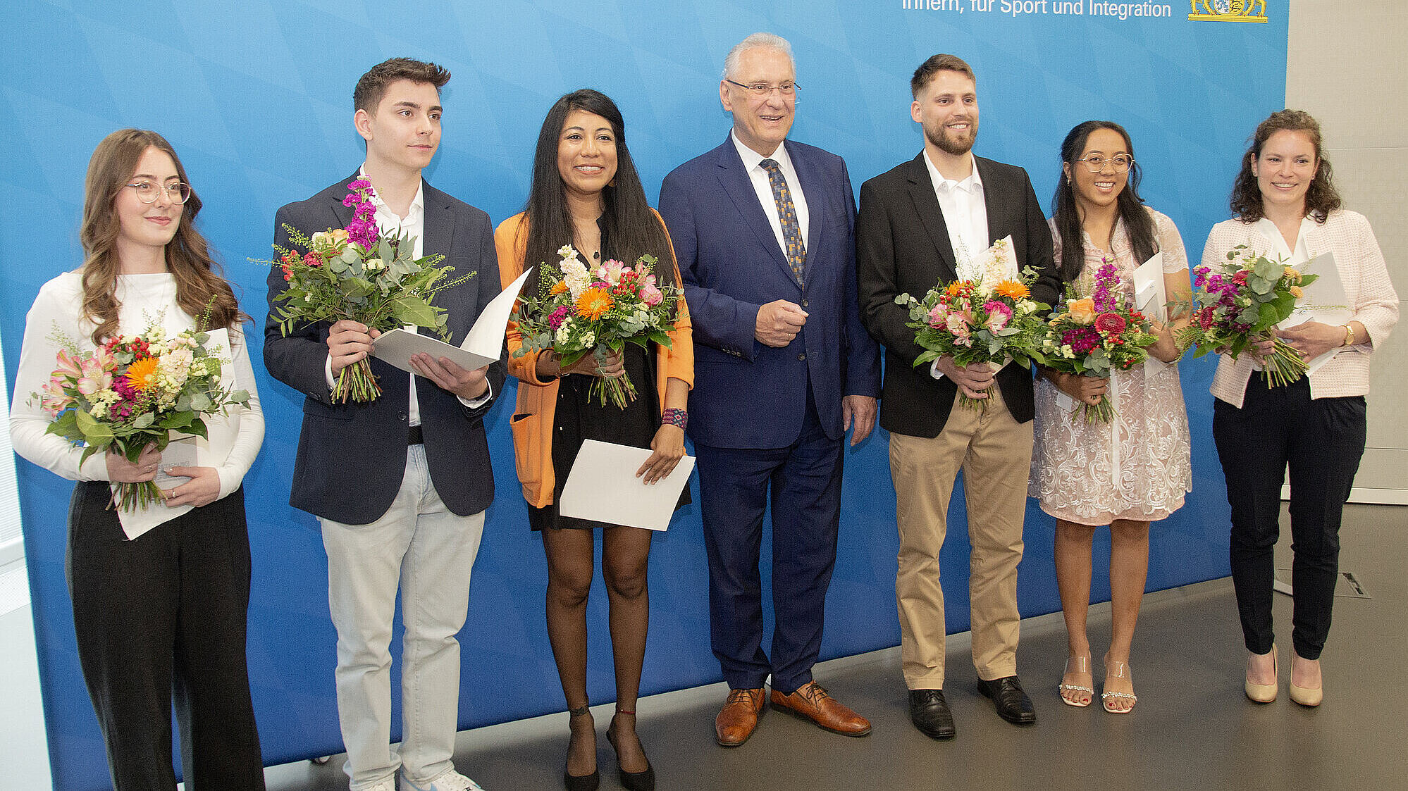Innenminister Herrmann zwischen sechs Personen mit Blumensträußen vor Pressewand