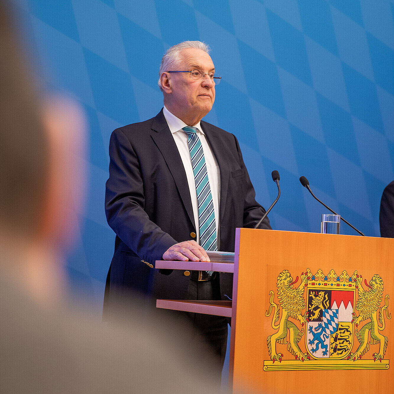 Innenminister Herrmann steht hinter Rednenrpult mit Bayerischem Staatswappen vor blauer Rautenwand 