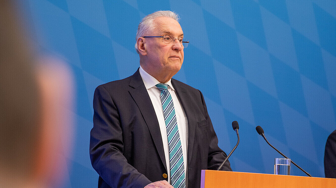 Innenminister Herrmann steht hinter Rednenrpult mit Bayerischem Staatswappen vor blauer Rautenwand 