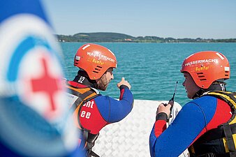 Zwei BRK-Ehrenamtliche besprechen sich über die Lage in Rettungsboot im Wasser, eine Person zeigt auf das entfernte Ufer