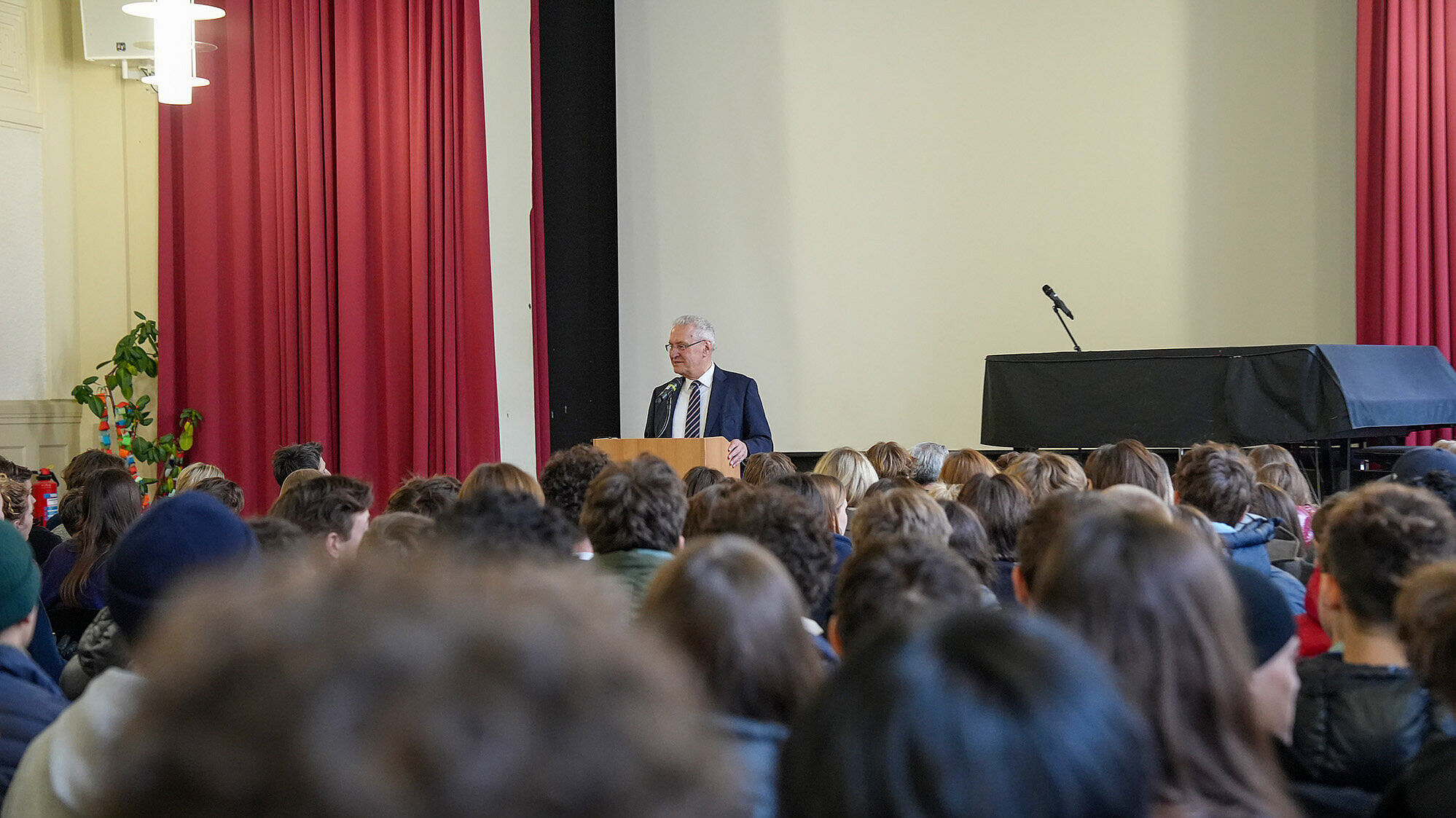 Innenminister Joachim Herrmann in Anzug und Krawatte steht an einem Rednerpult in einem großen Raum mit hohen Decken und roten Vorhängen. Vor ihm sitzt ein großes Publikum aus Jugendlichen und jungen Erwachsenen, die ihm zuhören. Rechts im Bild ist ein schwarzer Flügel zu sehen.