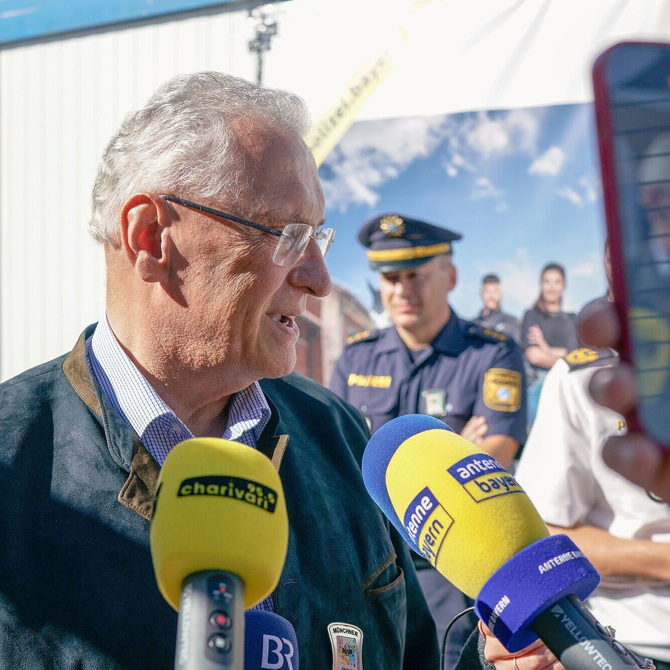 Auf dem Bild ist Innenminister Joachim Herrmann bei seinem Besuch der Wiesnwache 2024 zu sehen. Vor ihm sind mehrere Mikrofone zu sehen, in die er hineinspricht.