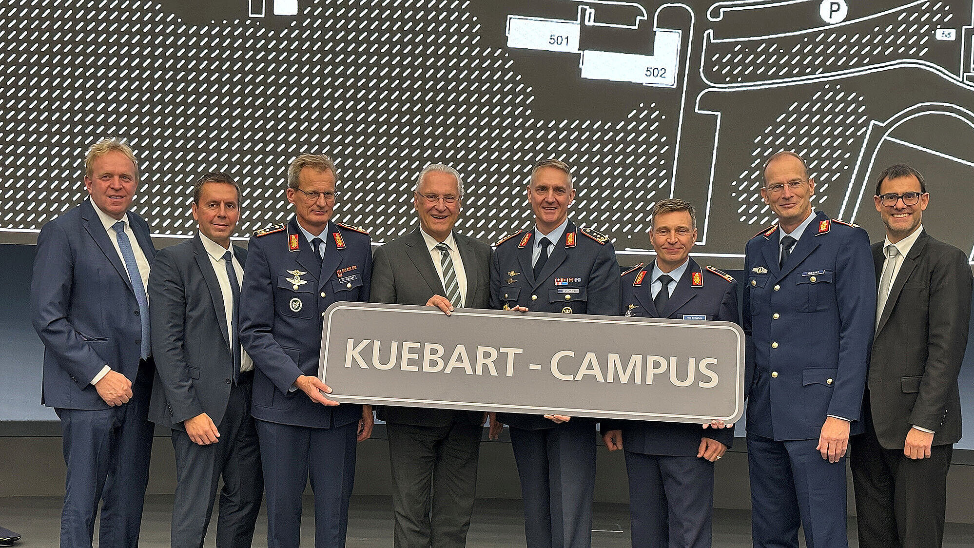 Auf dem Bild stehen acht Männer in formeller Kleidung (4. von li. Innenminister Joachim Herrmann), darunter mehrere in Uniformen der Luftwaffe mit Abzeichen und Orden. Sie halten gemeinsam ein großes Schild mit der Aufschrift „KUEBART-CAMPUS“. Im Hintergrund ist eine große Karte oder ein Plan an der Wand zu sehen.