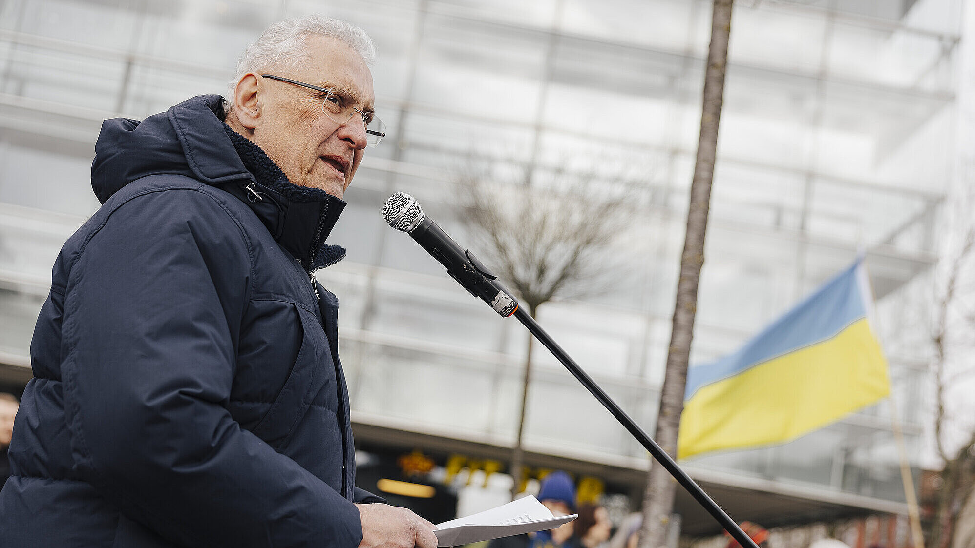 Person in dunkler Winterjacke spricht an einem Mikrofon und hält Papiere; im Hintergrund weht die ukrainische Flagge.