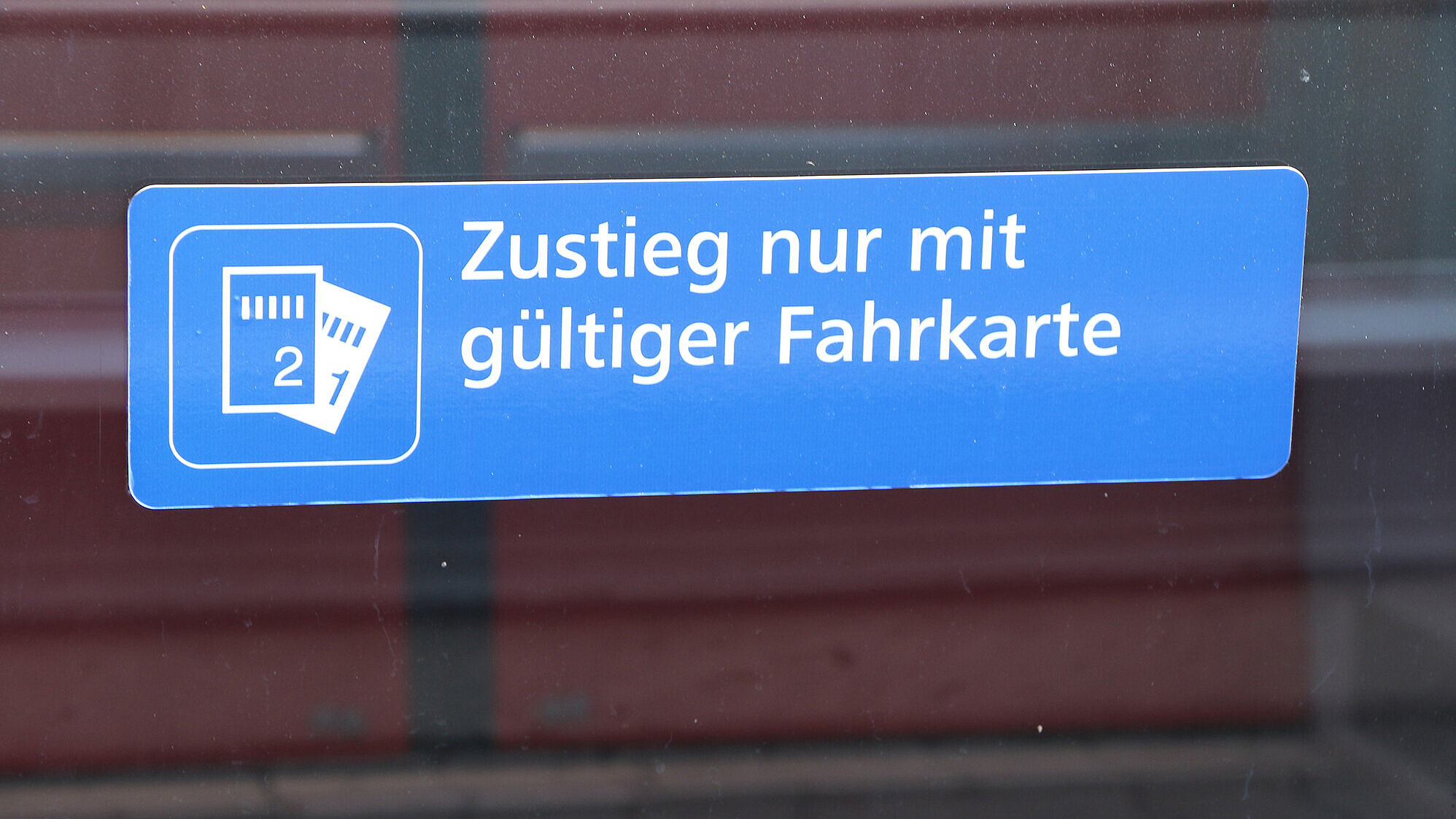 Blaues Schild an einer Fahrzeugtür mit der Aufschrift „Zustieg nur mit gültiger Fahrkarte“ und einem Piktogramm, das zwei Fahrkarten zeigt.
