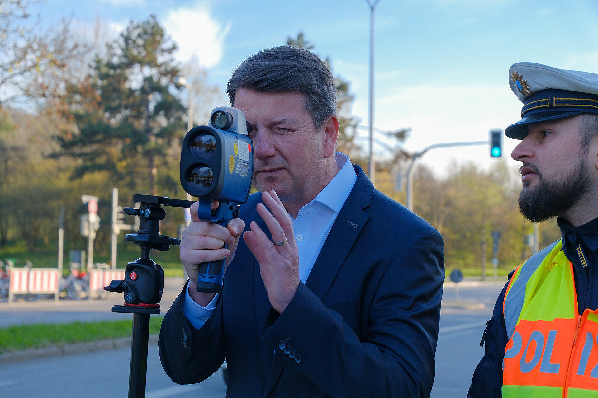 Person in Anzug blickt mit einem Auge durch ein Messgerät in seiner Hand, rechts angeschnitten ein Polizist in Uniform