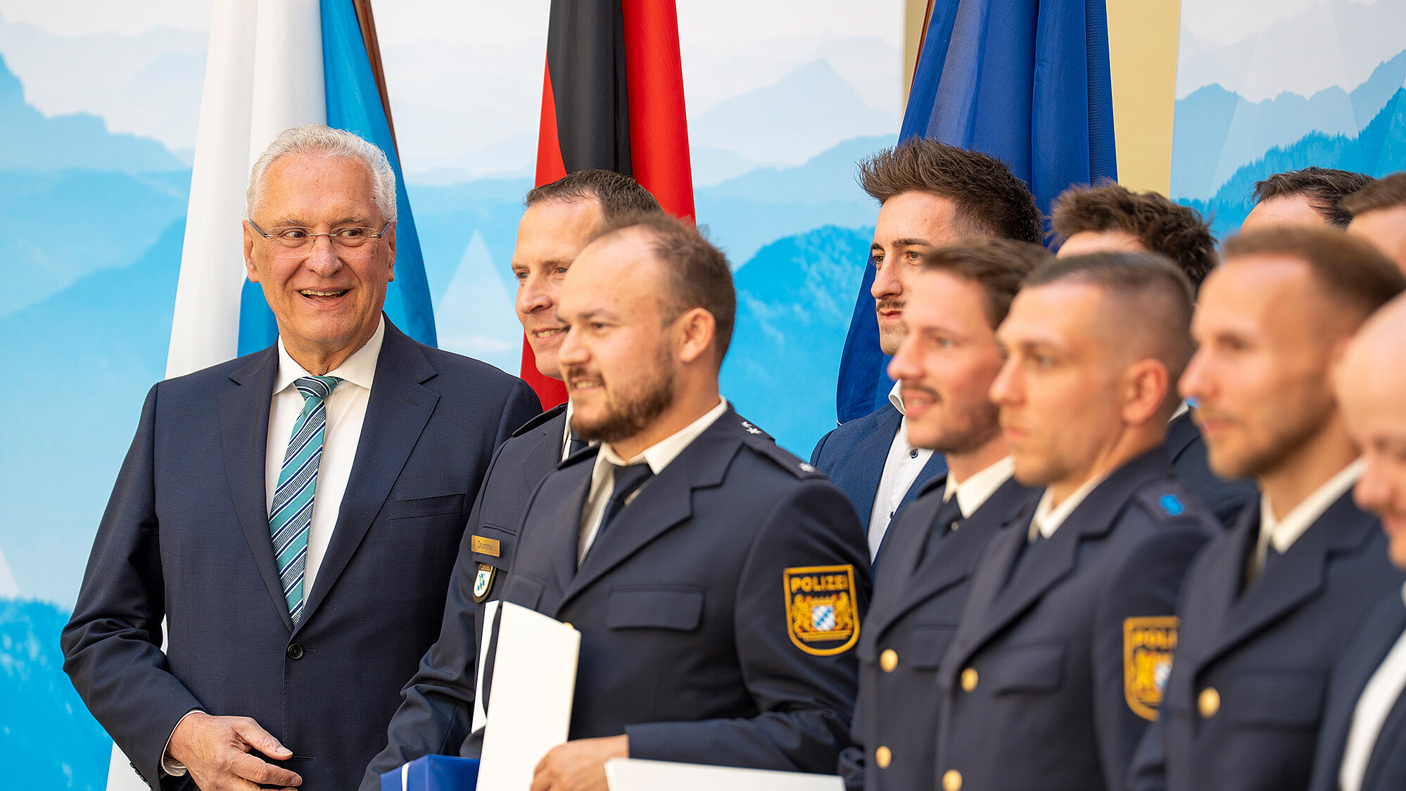 Mehrere Personen in Polizeiuniformen stehen nebeneinander bei einer Ehrungsveranstaltung; einige halten blau-weiße Geschenkboxen und Urkunden, im Hintergrund mehrere Flaggen und ein Bergmotiv; links ein Mann im Anzug