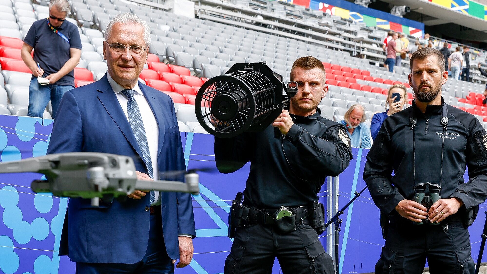 Herrmann neben zwei Polizisten, einer richtet Drohnenabwehrgerät auf Drohne, im Stadion