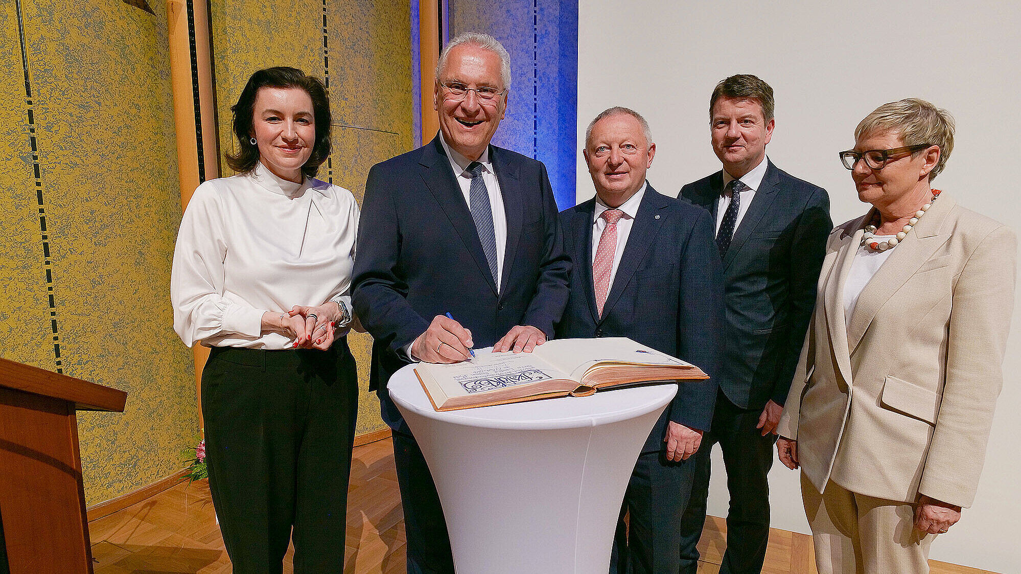 Fünf Personen in formeller Kleidung stehen um einen hohen weißen Stehtisch; eine Person unterschreibt ein aufgeschlagenes Gästebuch, die anderen blicken zu, Hintergrund: Saalwand mit Dekor.