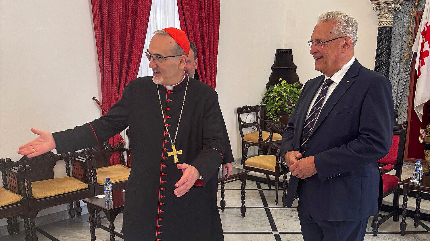 Kardinal Pizzaballa in traditioneller schwarzer Robe mit rotem Besatz und Kreuzkette begrüßt freundlich Innenminister Joachim Herrmann in einem festlich geschmückten Raum mit roten Vorhängen, Ikonen und kunstvoller Wandgestaltung.“
