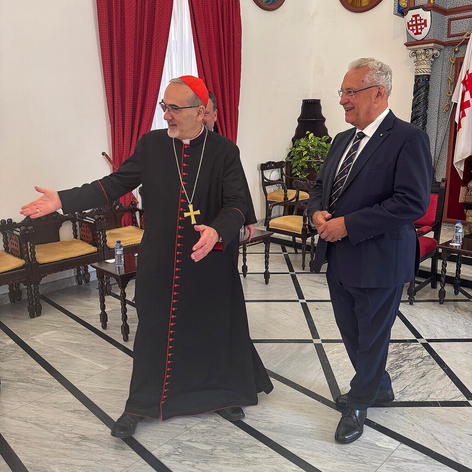 Kardinal Pizzaballa in traditioneller schwarzer Robe mit rotem Besatz und Kreuzkette begrüßt freundlich Innenminister Joachim Herrmann in einem festlich geschmückten Raum mit roten Vorhängen, Ikonen und kunstvoller Wandgestaltung.“