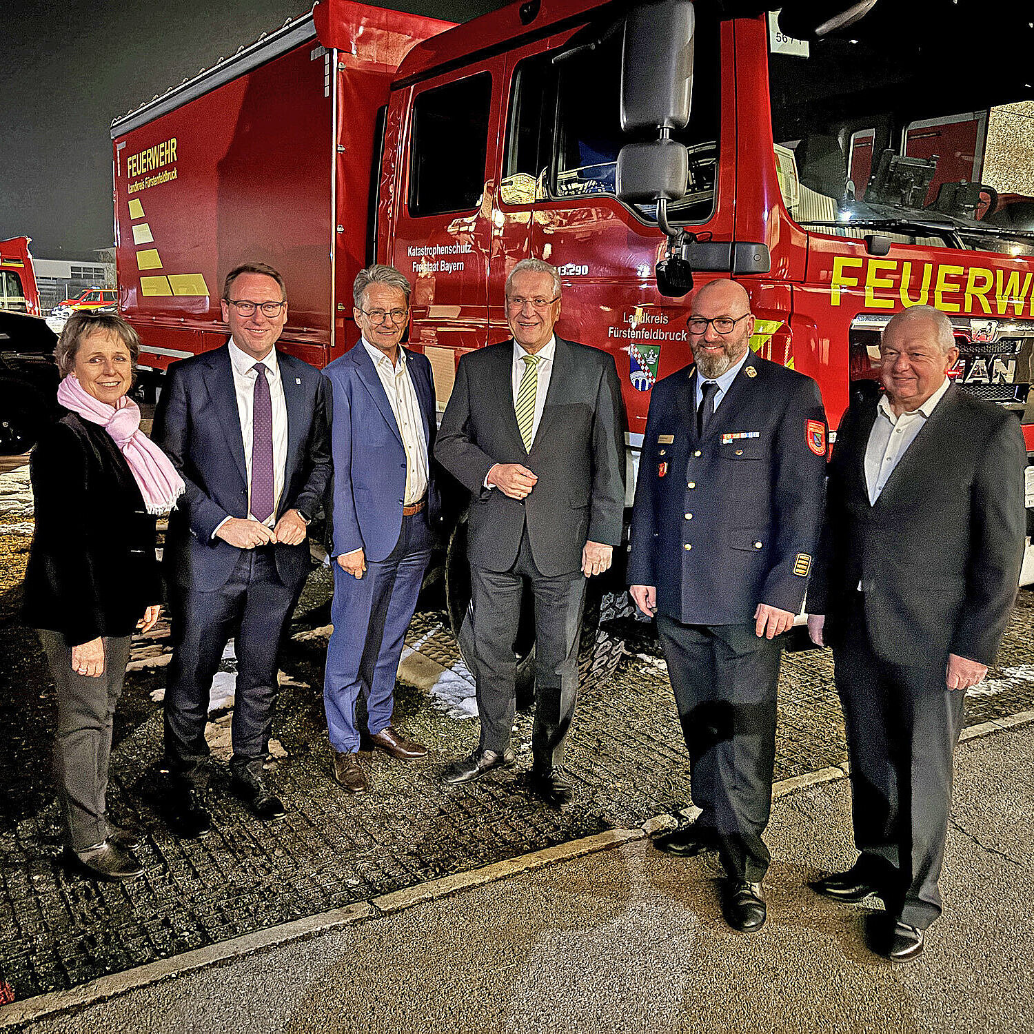 Sechs Personen stehen abends nebeneinander vor einem roten Feuerwehr-Lkw mit der großen Aufschrift FEUERWEHR; eine Person trägt Dienstuniform, die übrigen formelle Kleidung.