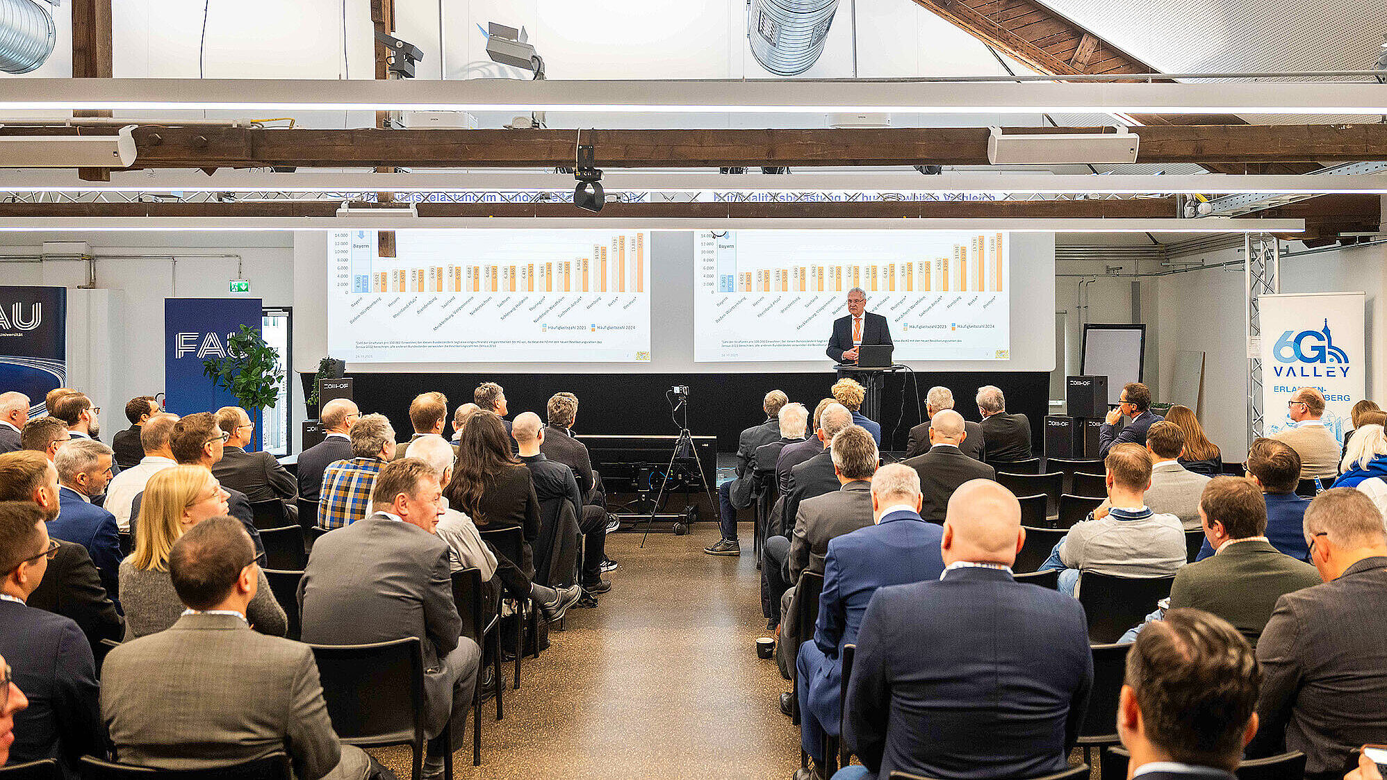 Ein Vortrag in einem modernen Konferenzraum mit vielen Sitzplätzen, die fast vollständig von einem Publikum in Businesskleidung besetzt sind. Der Redner ist Innenminister Joachim und steht vorne auf einer Bühne vor zwei großen Projektionsflächen, auf denen Diagramme mit Balkengrafiken zu sehen sind. Im Raum sind sichtbare Holzbalken an der Decke und technische Installationen. Auf der rechten Seite steht ein Banner mit dem Logo „6G Valley Erlangen Nürnberg“.