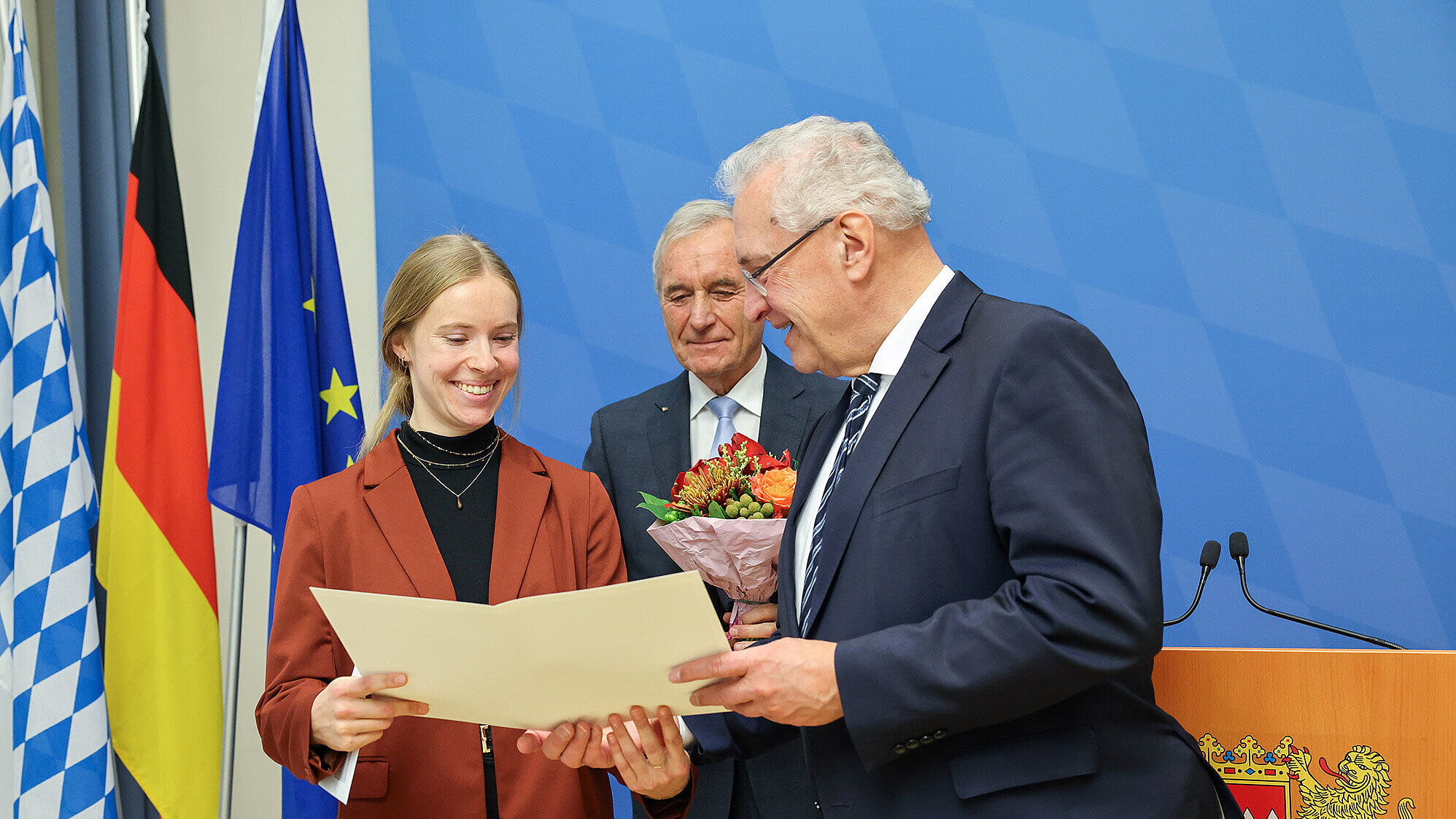 Eine Frau und zwei Männer (1.v.li. Innenminister Joachim Herrmann) in Anzügen stehen in einem Raum mit blauer Wand und bayerischer sowie europäischer Flagge im Hintergrund. Zwei von ihnen geben sich die Hand, während der dritte Mann ein geöffnetes Dokument hält und es zeigt. Die Szene wirkt formell und erinnert an eine Preisverleihung oder offizielle Übergabe.