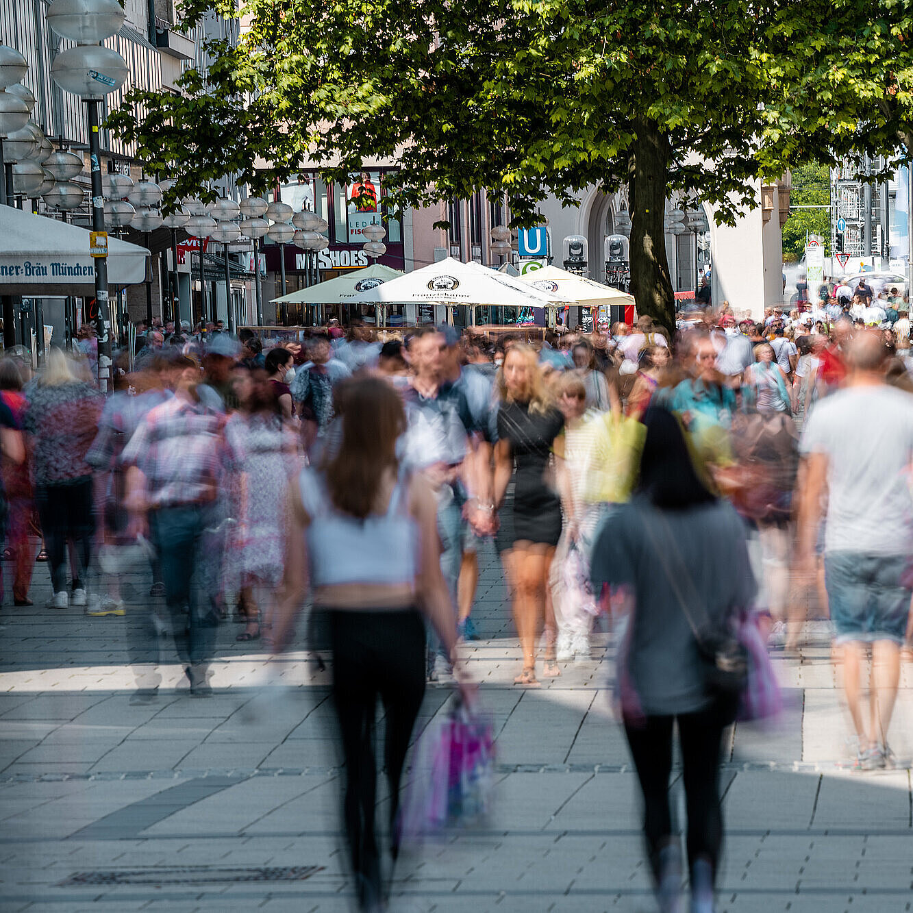 Passanten sind von vorne und hinten in der Münchner Fußgängerzone zu sehen. Im vorderen Bildbereich sind die Personen unscharf abgebildet.