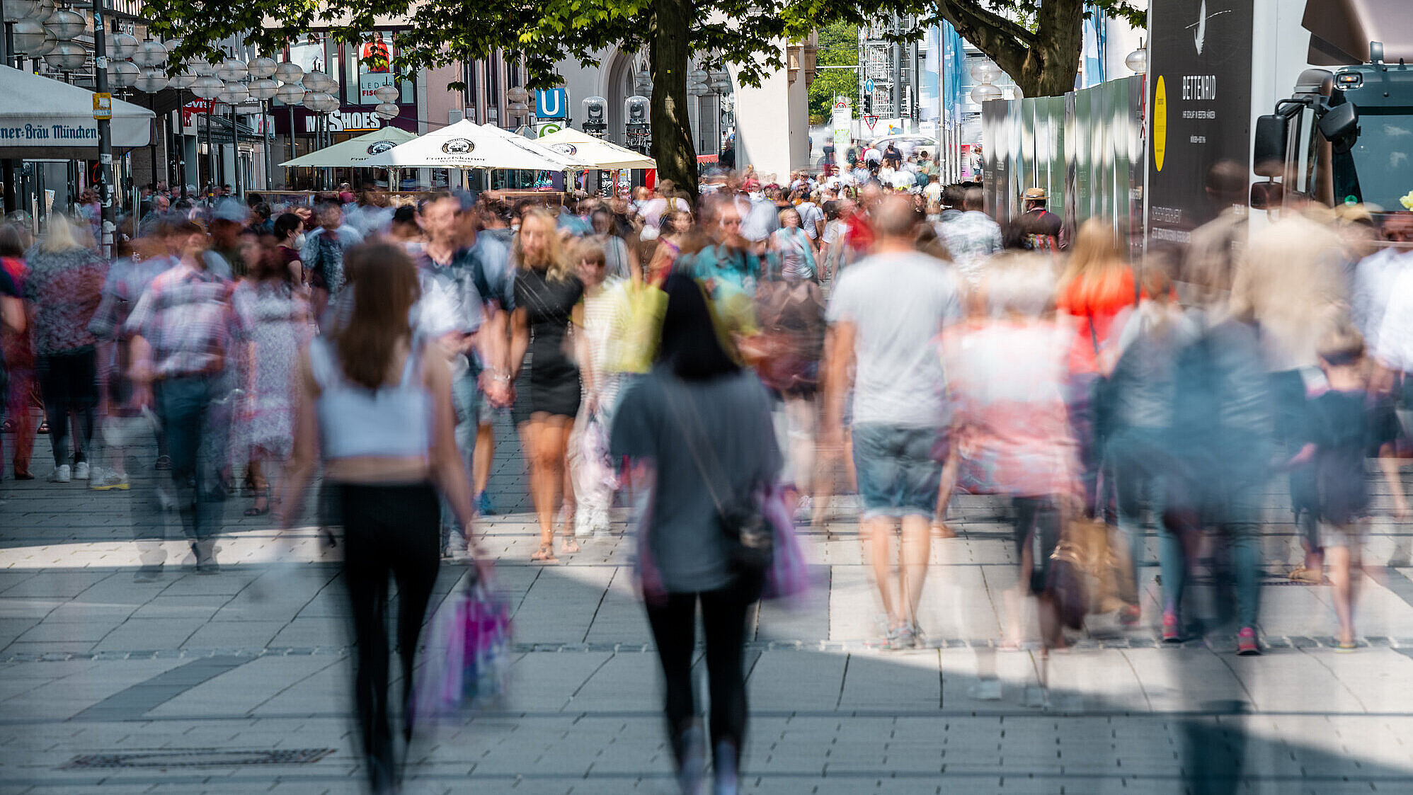 Passanten sind von vorne und hinten in der Münchner Fußgängerzone zu sehen. Im vorderen Bildbereich sind die Personen unscharf abgebildet.
