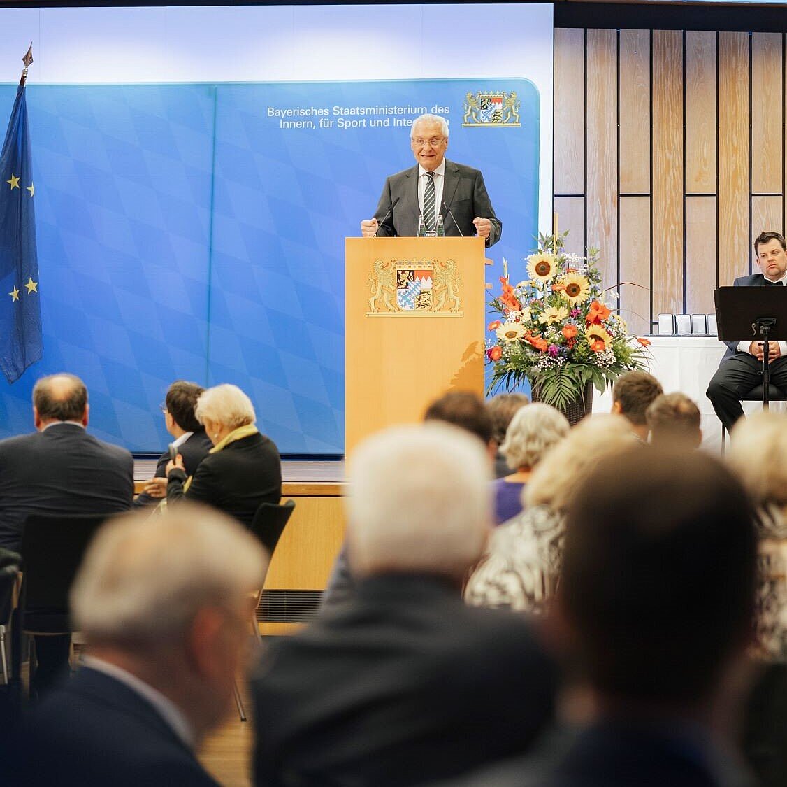 Auf dem Bild ist Innenminister Joachim Herrmann bei seiner Rede anlässlich der Ordensaushändigung am 30. Juli 2025 in Erlangen zu sehen. Vor ihm sieht man mehrere Gäste sitzen.