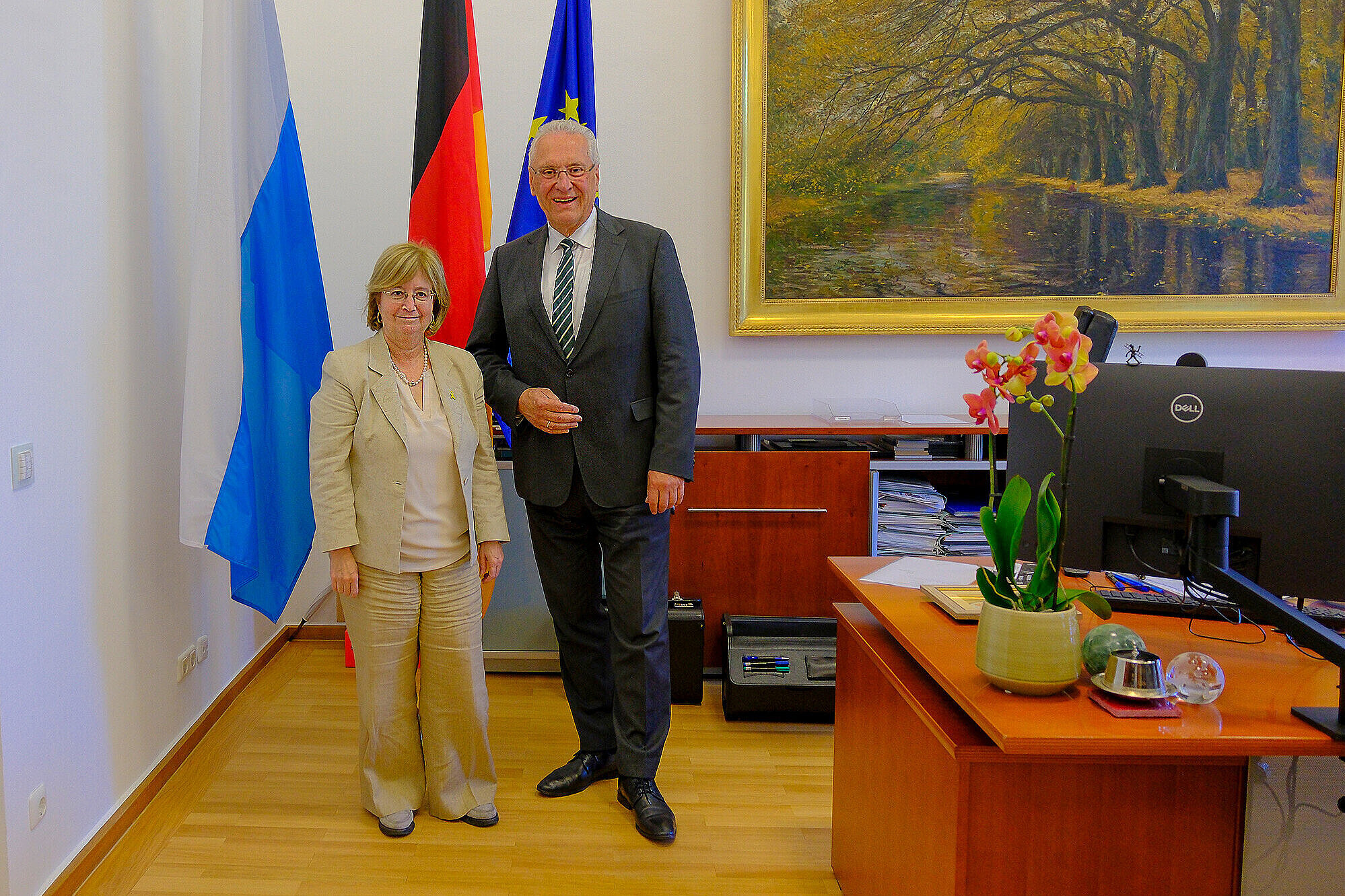 Innenminister Joachim Herrmann (re. im Bild) und die israelische Generalkonsulin Lador-Fresher stehen nebeneinander im Büro des Innenministers in München. Hinter ihnen sind die Flaggen von Deutschland, der Europäischen Union und einer weiteren Organisation zu sehen. Auf dem Tisch im Vordergrund stehen eine Orchidee in einer Vase, ein Computer, eine Lampe, eine Glaskugel und einige Unterlagen. An der Wand hängt ein großes Gemälde eines Flusses und eines Baumes im Herbst.