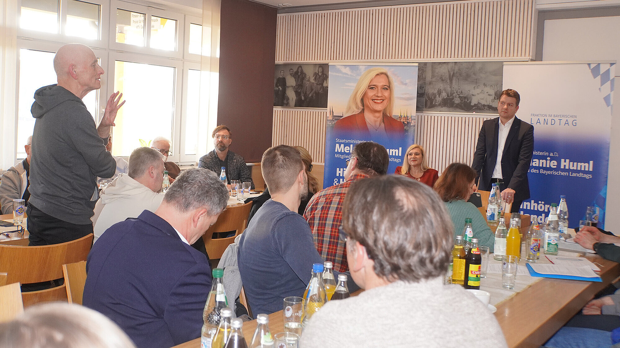 Mehrere Personen sitzen in einem hellen Raum an Tischen, auf denen verschiedene Getränke und Unterlagen stehen. Eine Person steht und spricht, während eine weitere Person vorne im Raum neben zwei großen Bannern steht. Die Banner zeigen Namen und Titel von Mitgliedern des Bayerischen Landtags. Im Hintergrund sind Fenster und Wandbilder zu sehen.