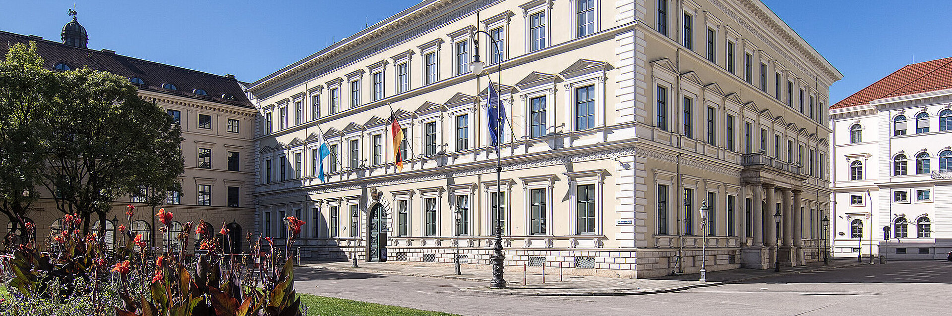 Gebäude des Bayerischen Innenministeriums von außen, am Eingang Flaggen, im Vordergrund bepflanzte Grünfläche