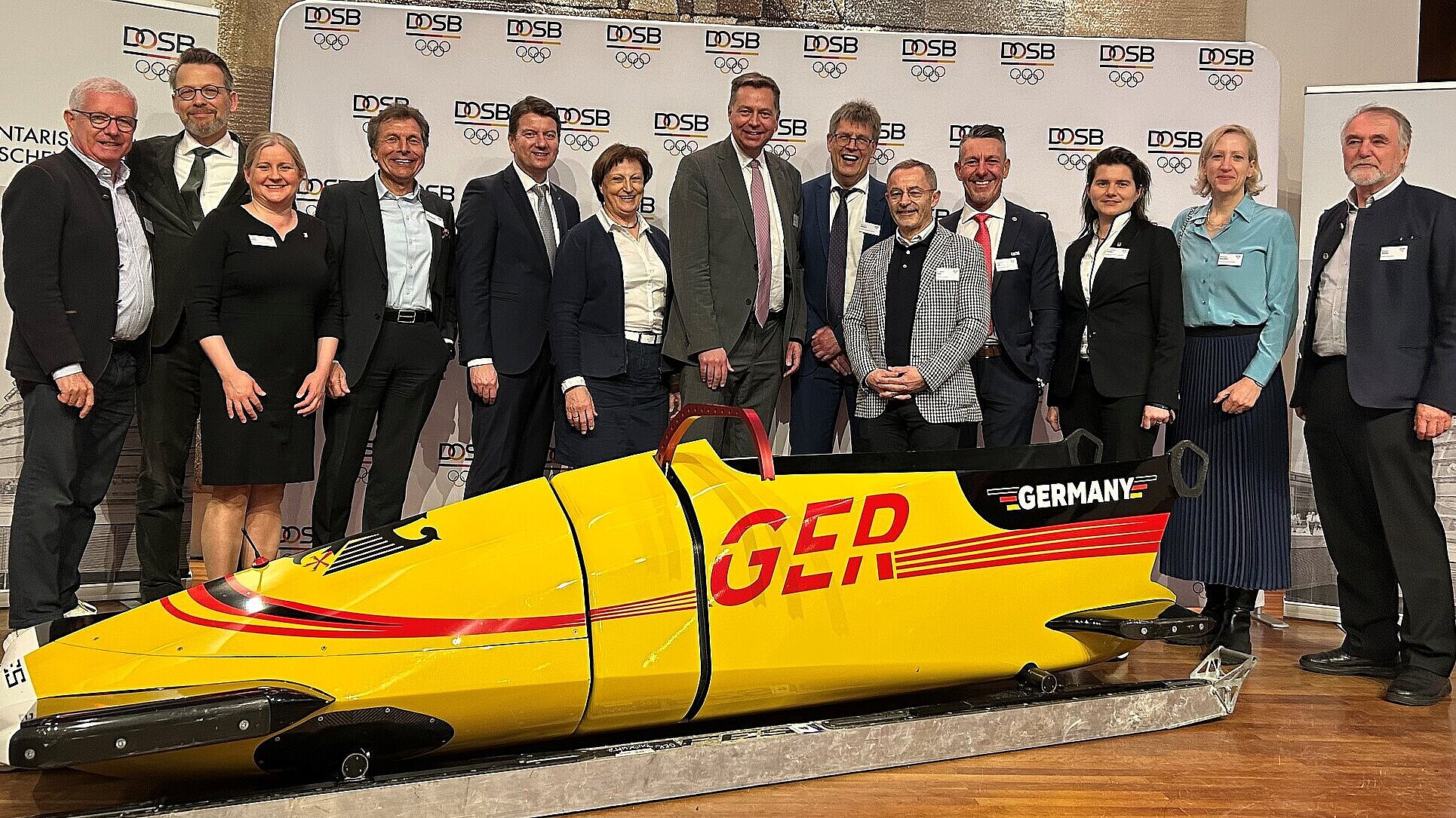 Auf dem Bild sind Staatssekretär Sandro Kirchner (5. v. l.) und verschiedene Politiker und Verbandsfunktionäre des DOSB zu sehen. Vor ihnen steht ein gelber Bob von Team Deutschland, hinter ihnen eine Pressewand des DOSB.