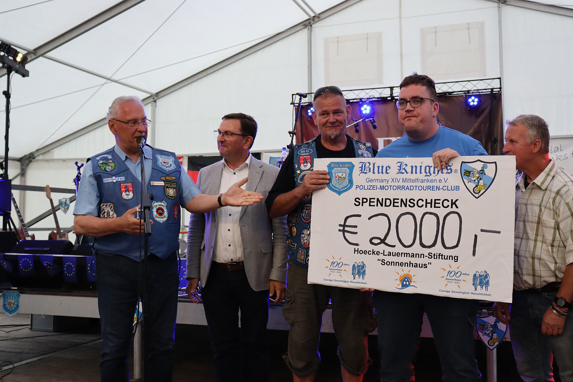 Innenminister Joachim Herrmann steht mit vier Mitgliedern der Blue Knights auf der Bühne und präsentiert Spendenscheck