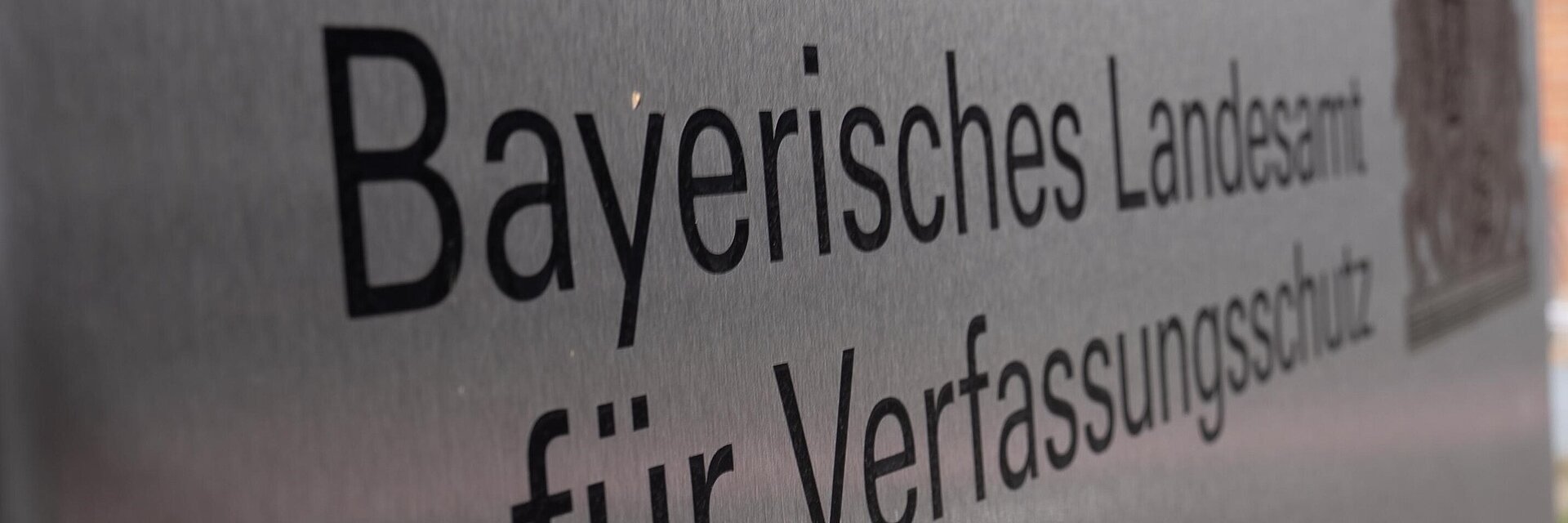 Schild mit Aufschrift Bayerisches Landesamt für Verfassungsschutz und dem Bayerischen Staatswappen schräg fotografiert