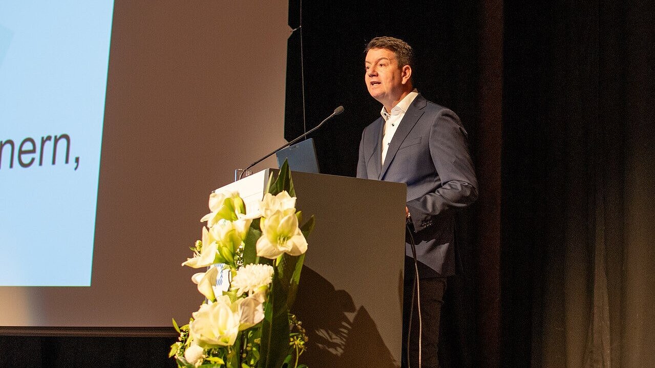 Innen- und Kommunalstaatssekretär Sandro Kircher auf der Bühne, hinter einem Rednerpult mit Blick ins Publikum bei seiner Festansprache vor ihm steht ein Blumengesteck mit weißen Lilien.