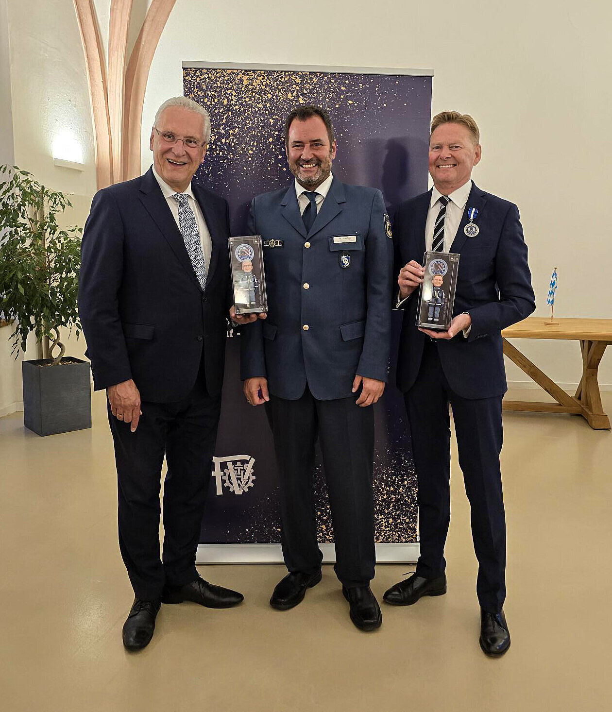 Innenminister Herrmann und zwei Personen vom THW in Anzug und Uniform mit verpackten Actionfiguren von sich selbst in der Hand
