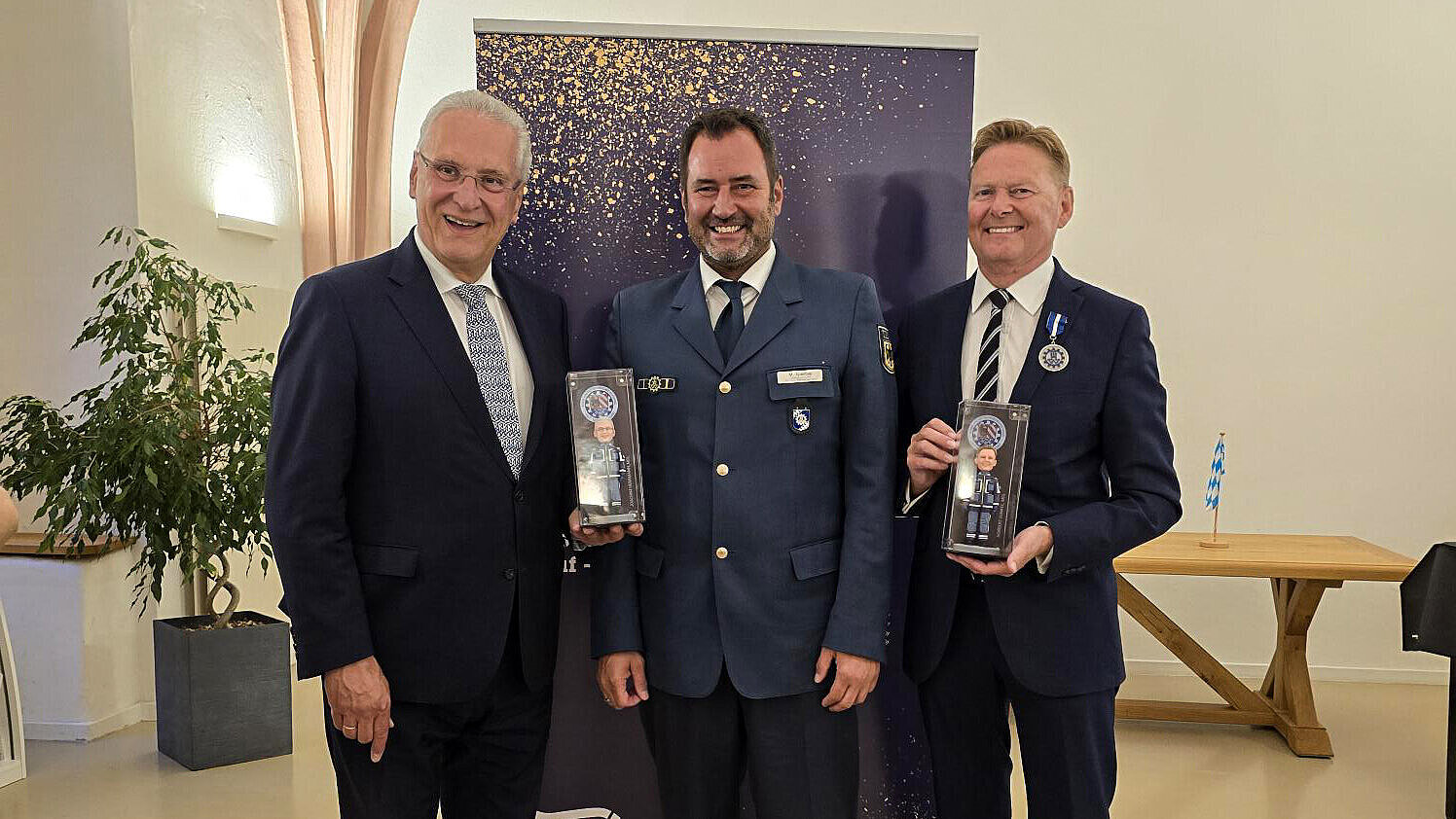 Innenminister Herrmann und zwei Personen vom THW in Anzug und Uniform mit verpackten Actionfiguren von sich selbst in der Hand