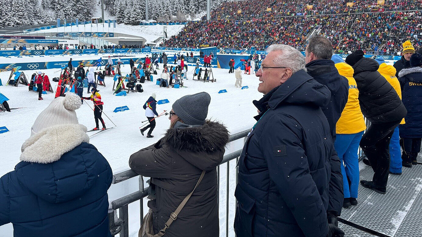 Zuschauer in Winterjacken lehnen an einer Absperrung und beobachten einen Skilanglauf‑Wettkampf auf verschneiter Strecke; Tribüne mit Zuschauern und Olympia‑Logos im Hintergrund.