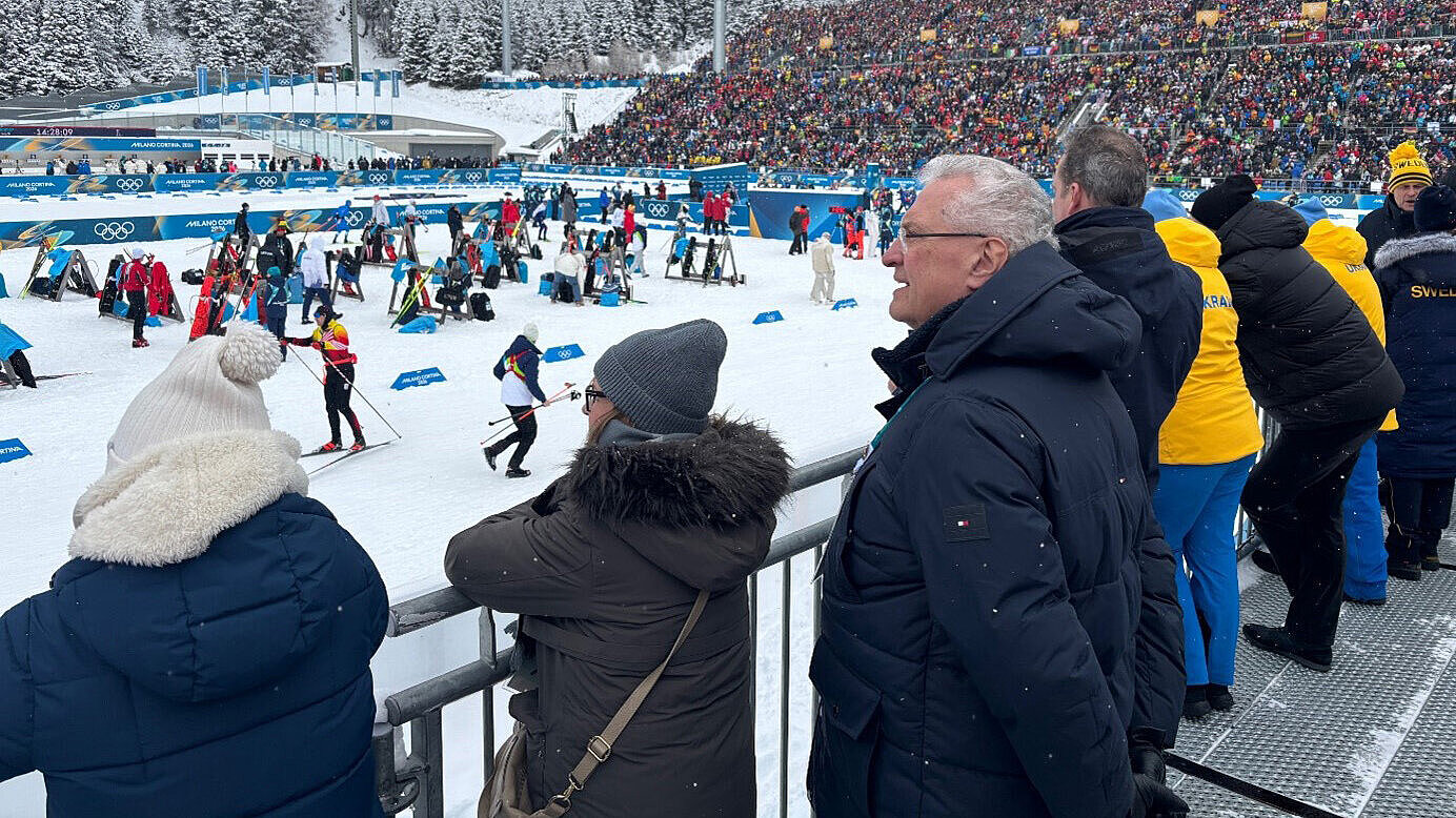 Zuschauer in Winterjacken lehnen an einer Absperrung und beobachten einen Skilanglauf‑Wettkampf auf verschneiter Strecke; Tribüne mit Zuschauern und Olympia‑Logos im Hintergrund.