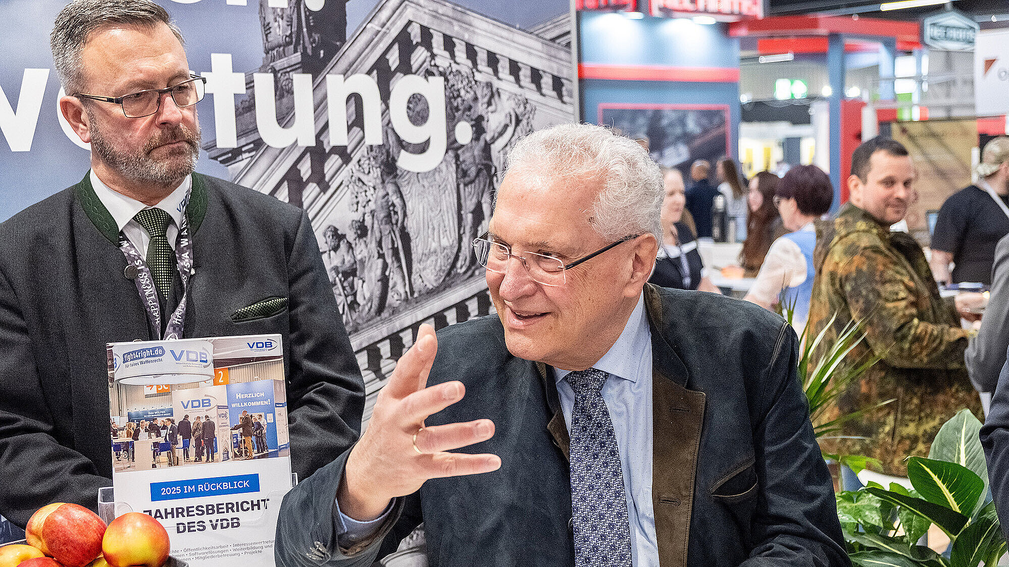 Zwei Männer sitzen an einem Messestand; einer spricht gestikulierend, im Hintergrund weitere Messebesucher und Pflanzen.