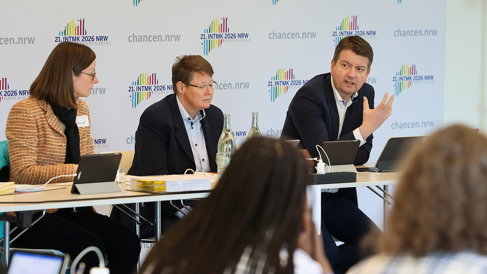 Konferenzszene: Drei Personen sitzen an einem Tisch vor einer Wand mit „21. INTMK 2026 NRW“ und „chancen.nrw“-Logos; im Vordergrund unscharfes Publikum, auf dem Tisch Tablets, Unterlagen und Wasserflaschen.