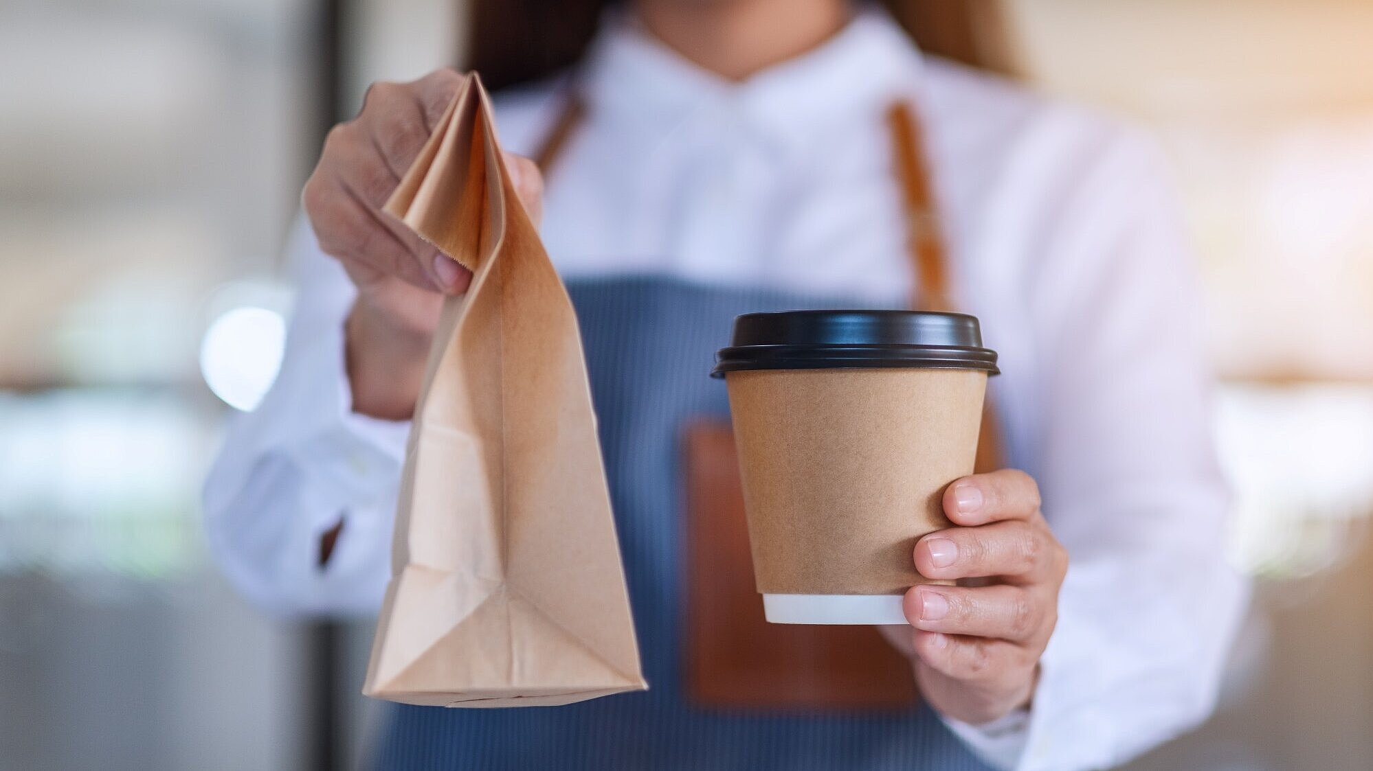Frau mit Schürze hält einen Coffee-to-go-Becher und eine Papiertüte