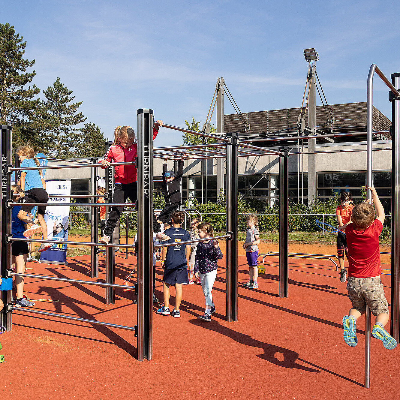 Kinder und Erwachsene nehmen an einer Sport- und Bewegungsveranstaltung im Freien teil. Auf einem roten Sportplatz nutzen die Kinder eine große Kletter- und Turnanlage mit verschiedenen Stangen und Sprossen. Erwachsene unterstützen oder beaufsichtigen die Kinder. Im Hintergrund sind Bäume, ein modernes Gebäude und Sportplatzbeleuchtung zu erkennen. Bei sonnigem Wetter herrscht eine freudige und aktive Atmosphäre.