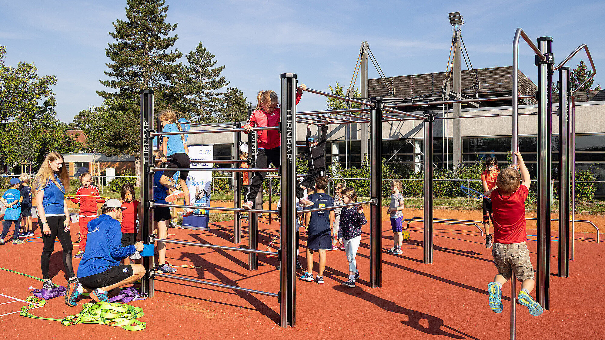 Kinder und Erwachsene nehmen an einer Sport- und Bewegungsveranstaltung im Freien teil. Auf einem roten Sportplatz nutzen die Kinder eine große Kletter- und Turnanlage mit verschiedenen Stangen und Sprossen. Erwachsene unterstützen oder beaufsichtigen die Kinder. Im Hintergrund sind Bäume, ein modernes Gebäude und Sportplatzbeleuchtung zu erkennen. Bei sonnigem Wetter herrscht eine freudige und aktive Atmosphäre.