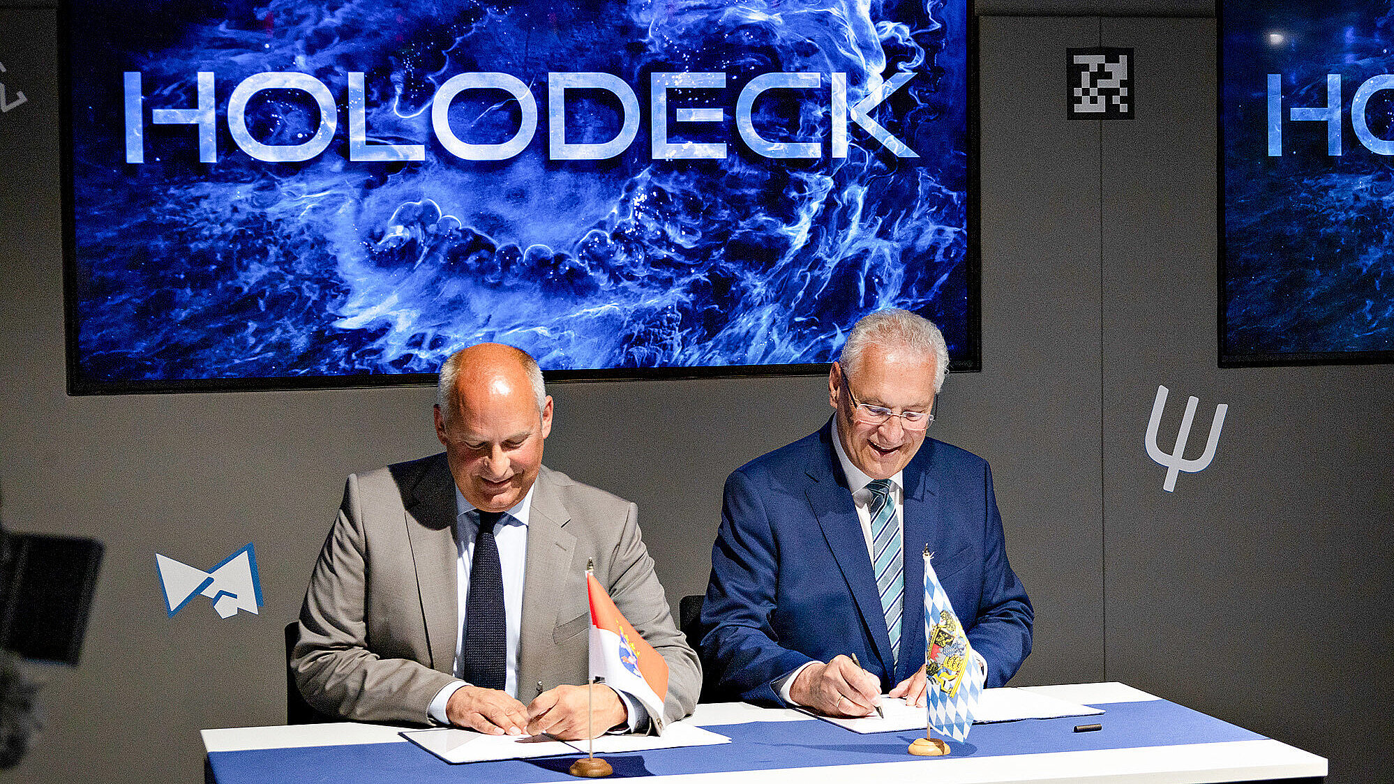 Zwei Männer in Anzügen sitzen an einem Tisch und unterschreiben Dokumente; im Hintergrund ein Bildschirm mit der Aufschrift „HOLODECK“.