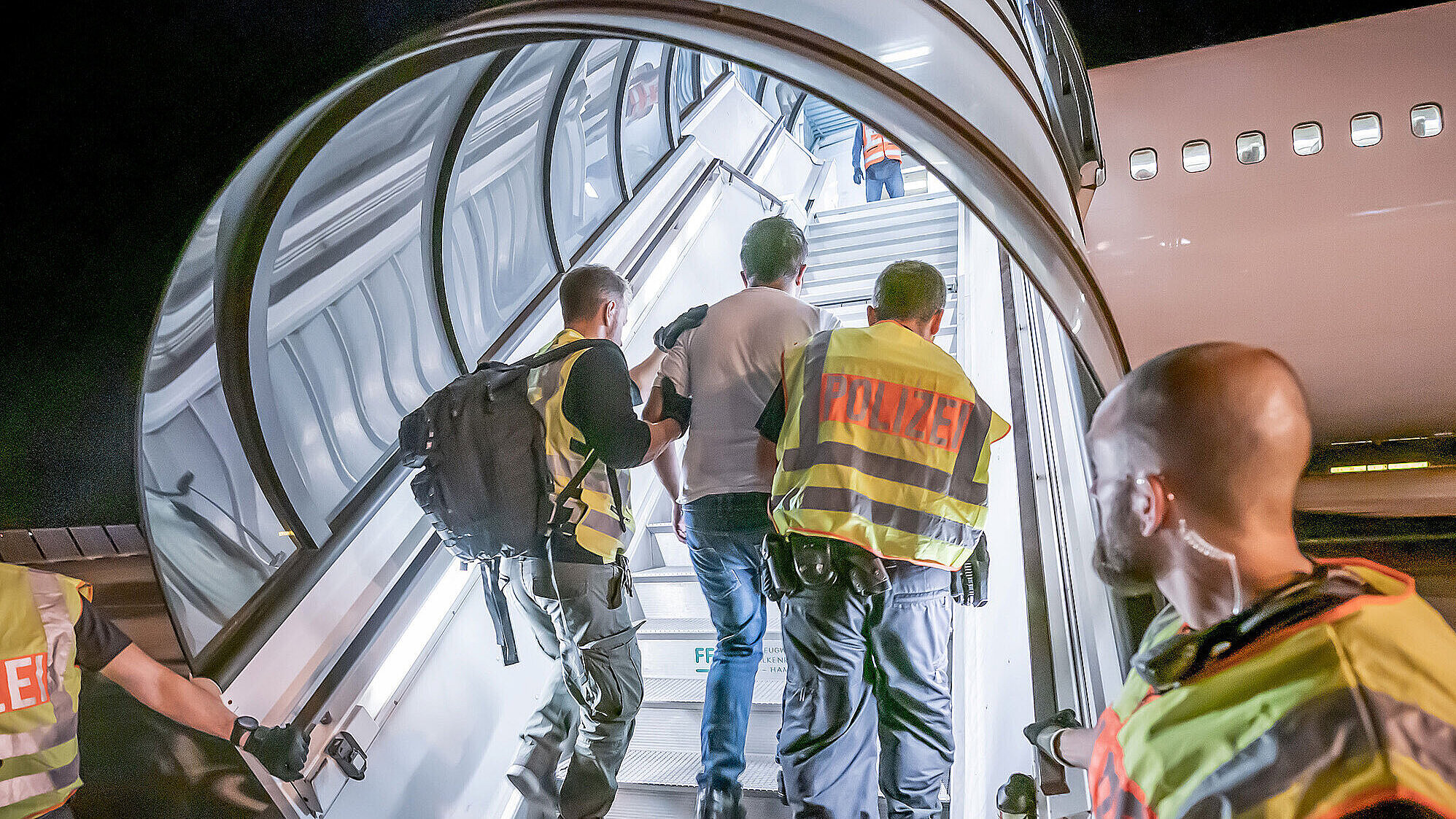 Mehrere Polizeibeamte begleiten bei Nacht einen abgelehnten Asylbewerber eine Flugzeugtreppe hinauf zu einem Abschiebeflug. Die Szene ist gut ausgeleuchtet, die Polizisten tragen Warnwesten mit der Aufschrift „Polizei“.