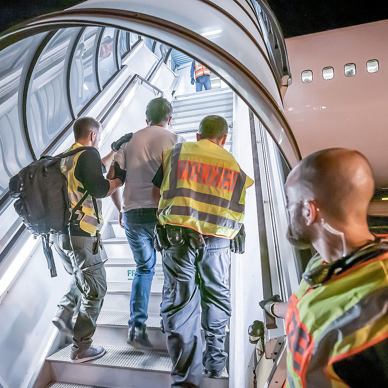 Mehrere Polizeibeamte begleiten bei Nacht einen abgelehnten Asylbewerber eine Flugzeugtreppe hinauf zu einem Abschiebeflug. Die Szene ist gut ausgeleuchtet, die Polizisten tragen Warnwesten mit der Aufschrift „Polizei“.