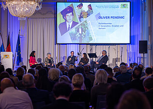 Herrmann und weitere Teilnehmerinnen und Teilnehmer sitzend auf der Bühne bei einer Podiumsdiskussion, im Hintergrund großer Bildschirm mit Präsentation, im Vordergrund Publikum von hinten.