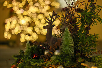 Weihnachtliche Tischdekoration mit Hirschfiguren, Tannenzweigen, Schmuckkugeln und Mini-Tännchen; im Hintergrund unscharfer, warm leuchtender Weihnachtsbaum mit Lichterkette.