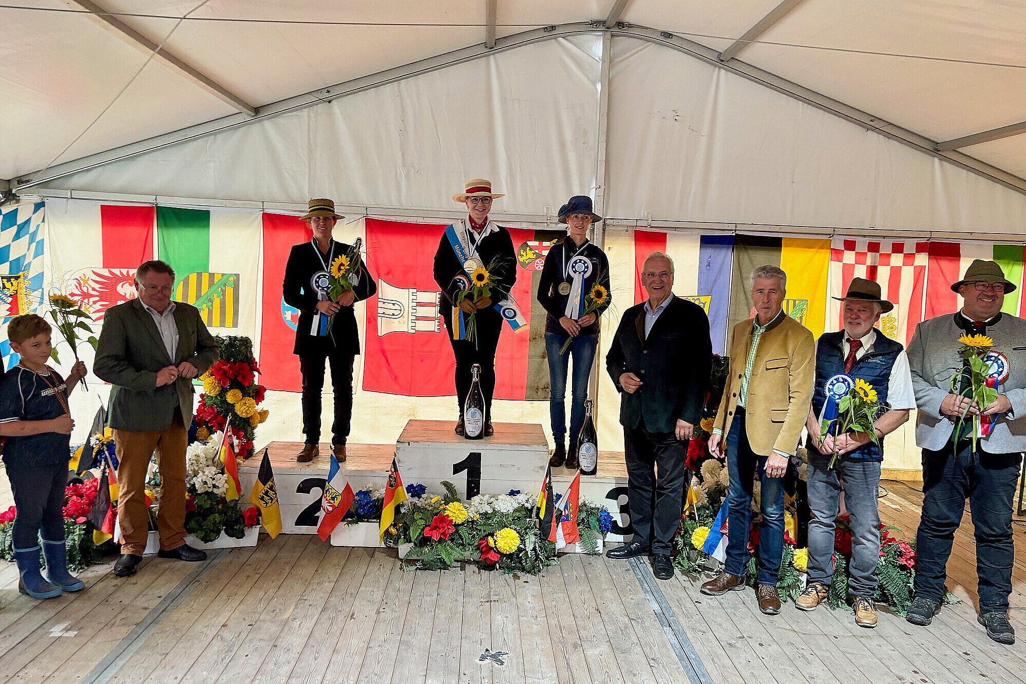 Siegerehrung beim Internationalen Fahrturnier unter einem Festzelt: Die Erstplatzierten stehen auf einem Podest und halten Schleifen, Blumen und Urkunden in den Händen. Hinter ihnen hängen zahlreiche Landesflaggen. Innenminister Joachim Herrmann, Gerhard Eck, Präsident des Reit- und Fahrverbandes und weitere Ehrengäste stehen daneben und gratulieren. Das Podest ist mit Blumen und kleinen Landesflaggen dekoriert.