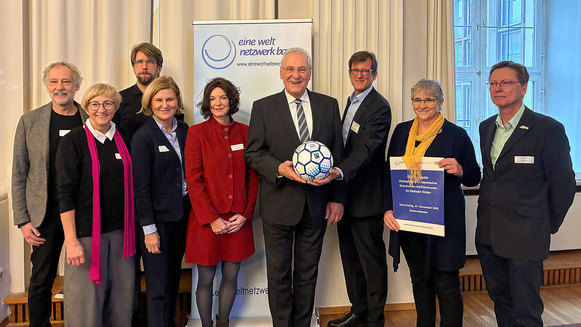 Gruppenfoto von neun Personen (4.v.li. Innenminister Joachim Herrmann), die in einem Raum mit hohen Fenstern und hellen Vorhängen stehen. In der Bildmitte hält eine Person in einem dunklen Anzug einen Fußball mit blauem Muster. Eine weitere Person hält ein Plakat mit Text. Im Hintergrund ist ein Roll-up mit dem Logo und Schriftzug des Eine-Welt-Netzwerks Bayern zu sehen. Alle Personen tragen Namensschilder.