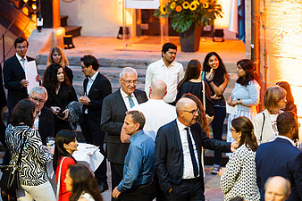 Zahlreiche Gäste im Gespräch bei einem Empfang im Innenhof der Kaiserburg Nürnberg. In der Mitte des Bildes unterhält sich Bayerns Innenminister Joachim Herrmann in einem dunklen Anzug mit weiteren Teilnehmenden. Im Hintergrund leuchten warme Lichter und eine große Blumendekoration mit Sonnenblumen sorgt für festliches Ambiente.