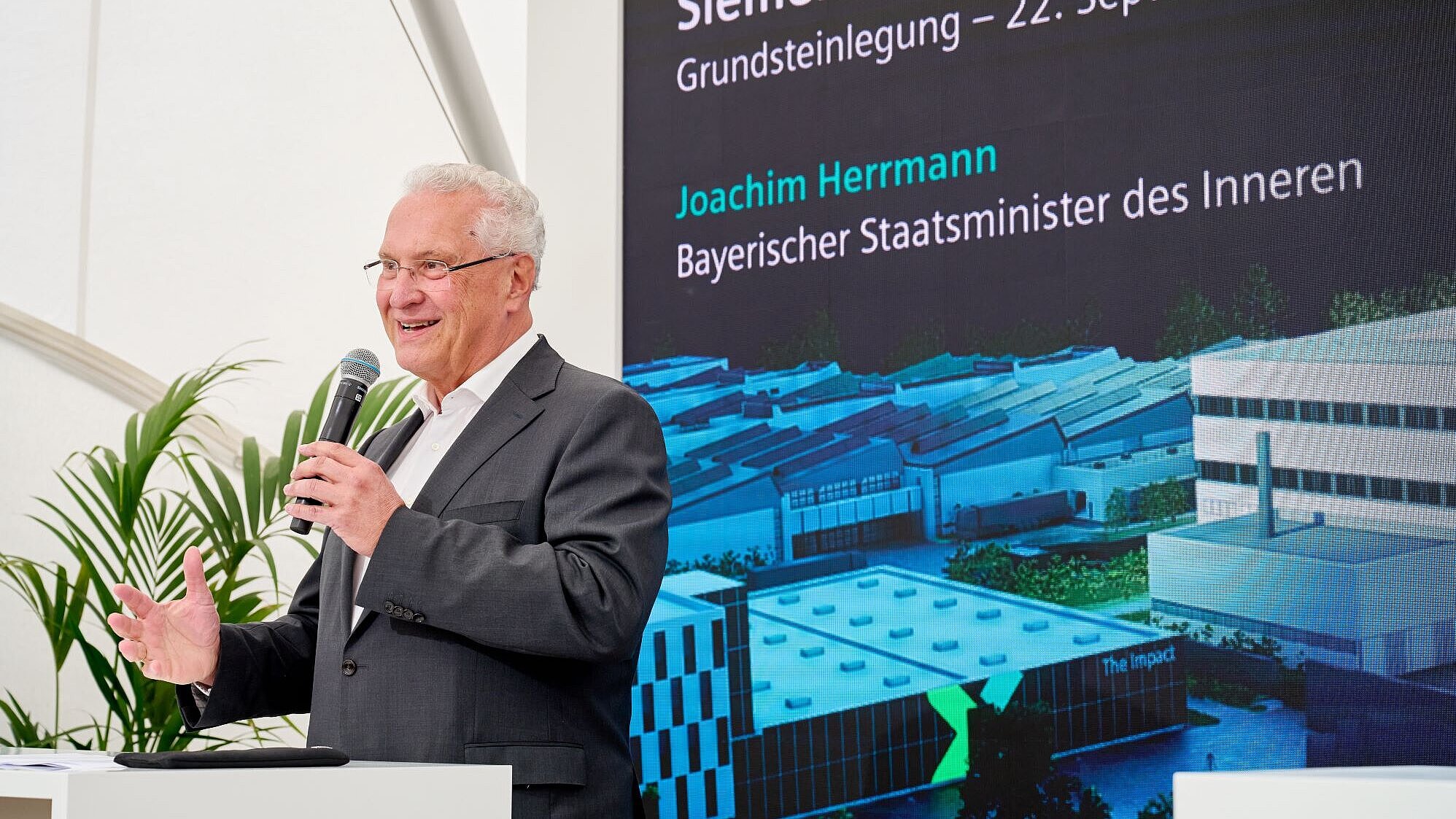 Joachim Herrmann ist hier bei seiner Rede anlässlich der Grundsteinlegung bei Siemens in Erlangen zu sehen. Hinter ihm sieht man eine Pressewand mit den Daten der Veranstaltung, Herrmann hält ein Mikrofon und lächelt ins Publikum.