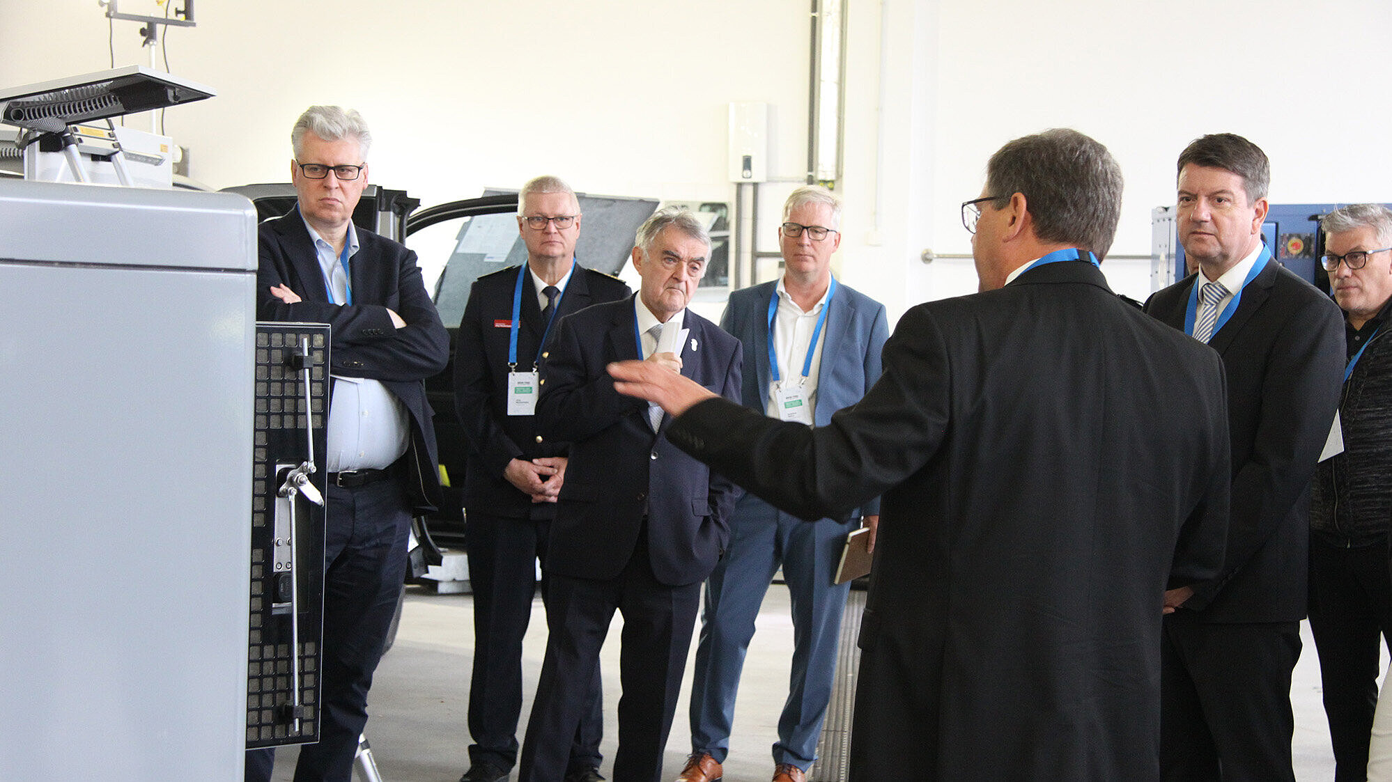 Das Bild zeigt eine Gruppe von Menschen, die in einem Raum stehen und einem Mann in einem schwarzen Anzug zuhören, der anscheinend eine Präsentation oder eine Erklärung gibt. Im Vordergrund ist eine Maschine oder ein technisches Gerät zu sehen. Die Personen tragen formelle Kleidung, beim BOS-Tag in Essen.