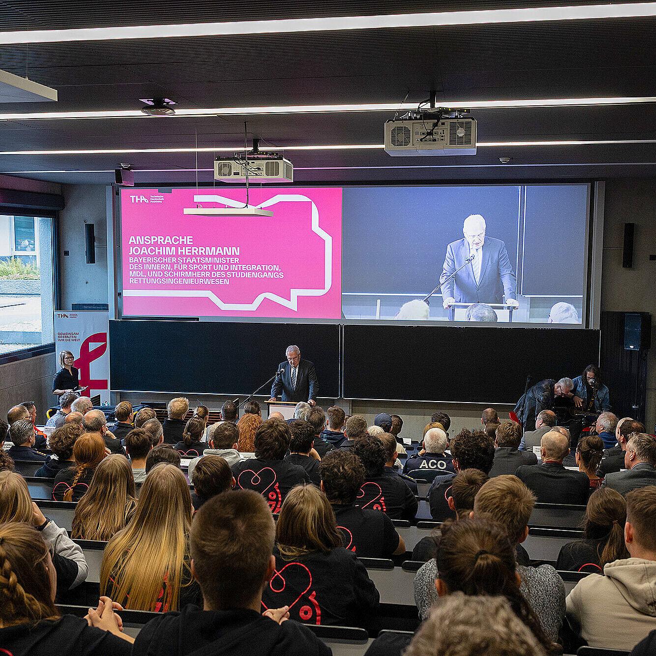 Ein Bild von einer Veranstaltung im Hörsaal, bei der Innenminister Joachim Herrmann am Rednerpult spricht. Auf der großen Leinwand im Hintergrund ist eine Präsentation mit dem Titel "Ansprache Joachim Herrmann" zu sehen. Der Saal ist voll mit Zuhörern, die aufmerksam dem Vortrag folgen.